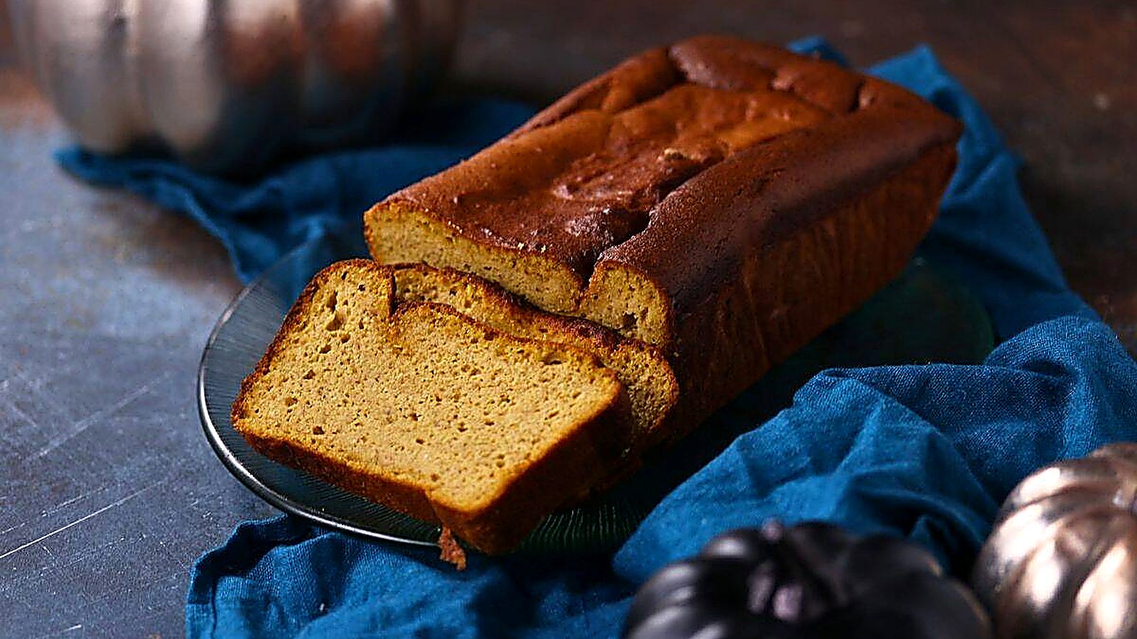 A loaf of pumpkin pound cake with two slices cut sits on a glass plate atop a blue cloth, with decorative pumpkins nearby.
