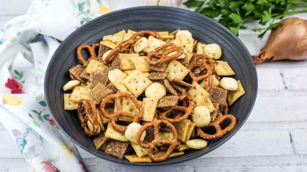 A black bowl filled with a snack mix of square crackers, pretzels, cereal pieces, and round crackers, placed on a light surface near a floral napkin and fresh herbs.