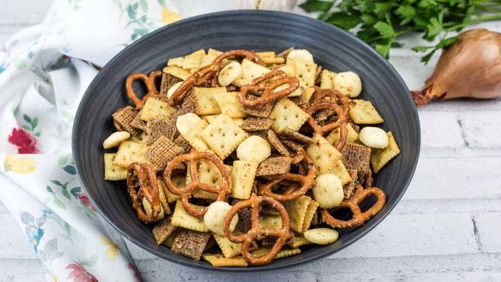 A black bowl filled with snack mix including pretzels, square crackers, cereal squares, and round crackers, sits on a white surface next to a floral napkin, parsley, and a shallot.