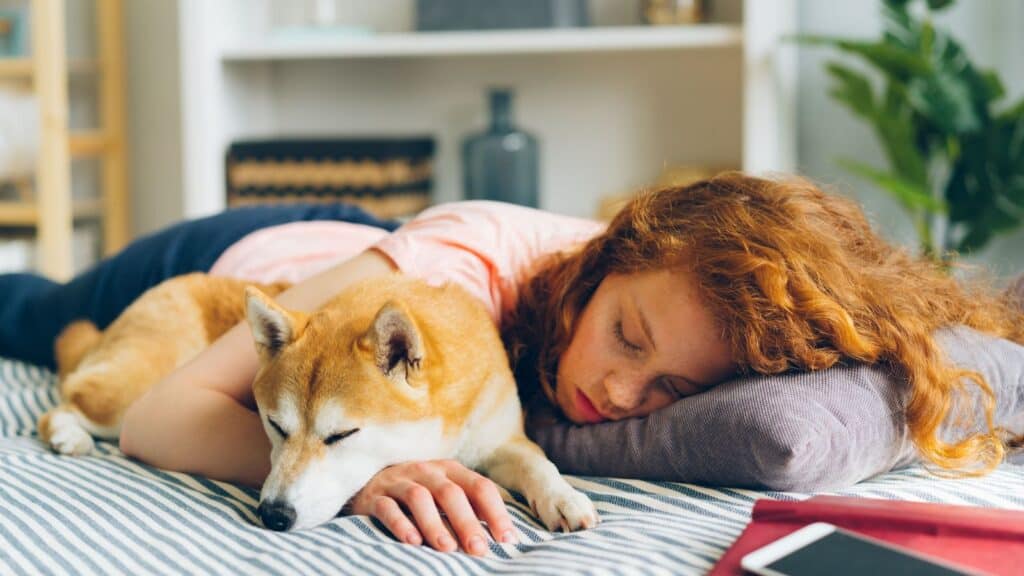 A young woman with red hair is lying on a bed, resting her head on a pillow next to a Shiba Inu dog. Both appear to be asleep, embodying the perfect scene of jet lag recovery as recommended by sleep science experts.