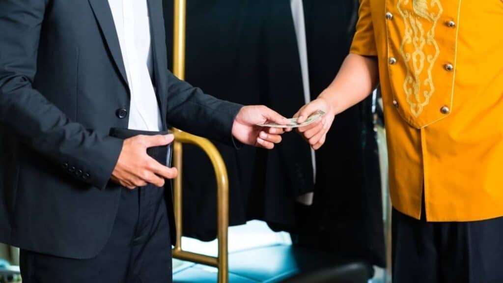 A person in a suit hands money to a person in a yellow uniform, standing near a luggage cart, possibly discussing the all inclusive resort fine print before their stay begins.