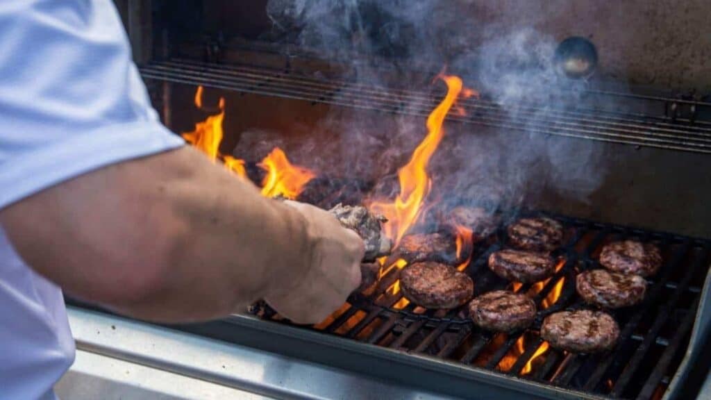 A person grilling hamburger patties on a barbecue with visible flames and smoke rising from the grill, using tailgating gear from a luxury category.