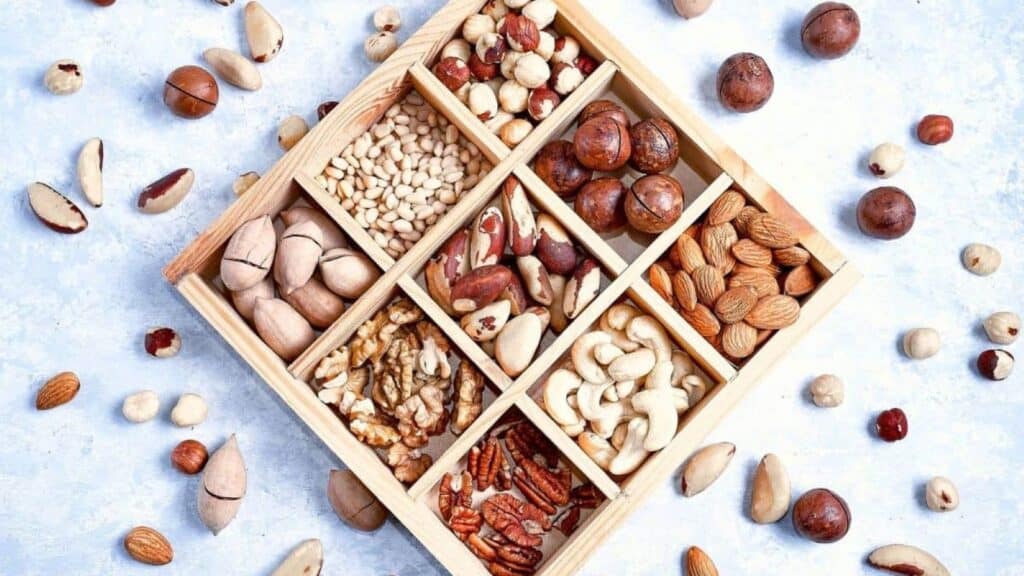 A wooden box with nine sections, each filled with different types of nuts&mdash;a perfect choice among college care package items&mdash;surrounded by assorted loose nuts on a light surface.