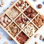 A wooden box with nine sections, each filled with different types of nuts—a perfect choice among college care package items—surrounded by assorted loose nuts on a light surface.