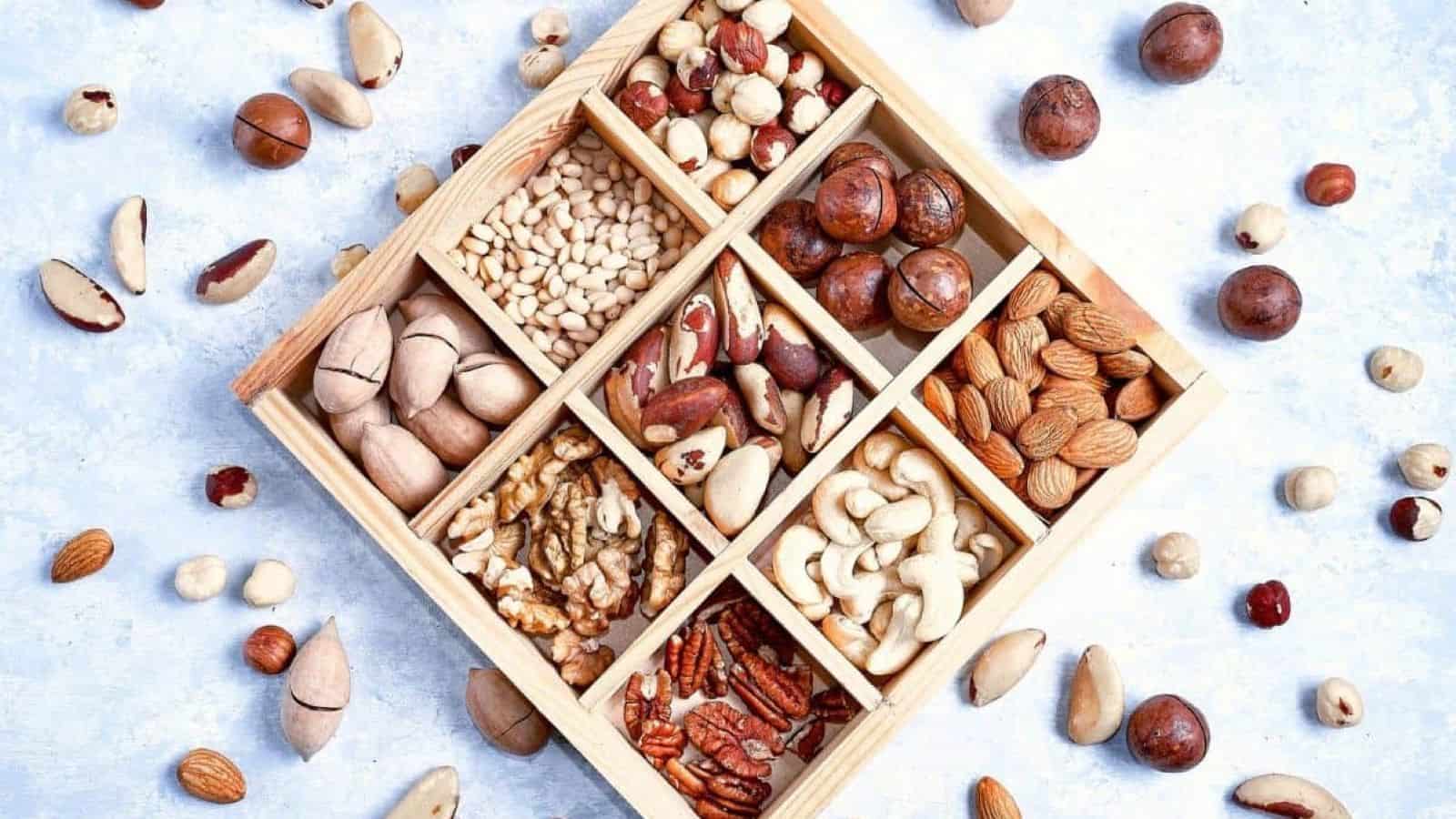 A wooden box with nine sections, each filled with different types of nuts—a perfect choice among college care package items—surrounded by assorted loose nuts on a light surface.