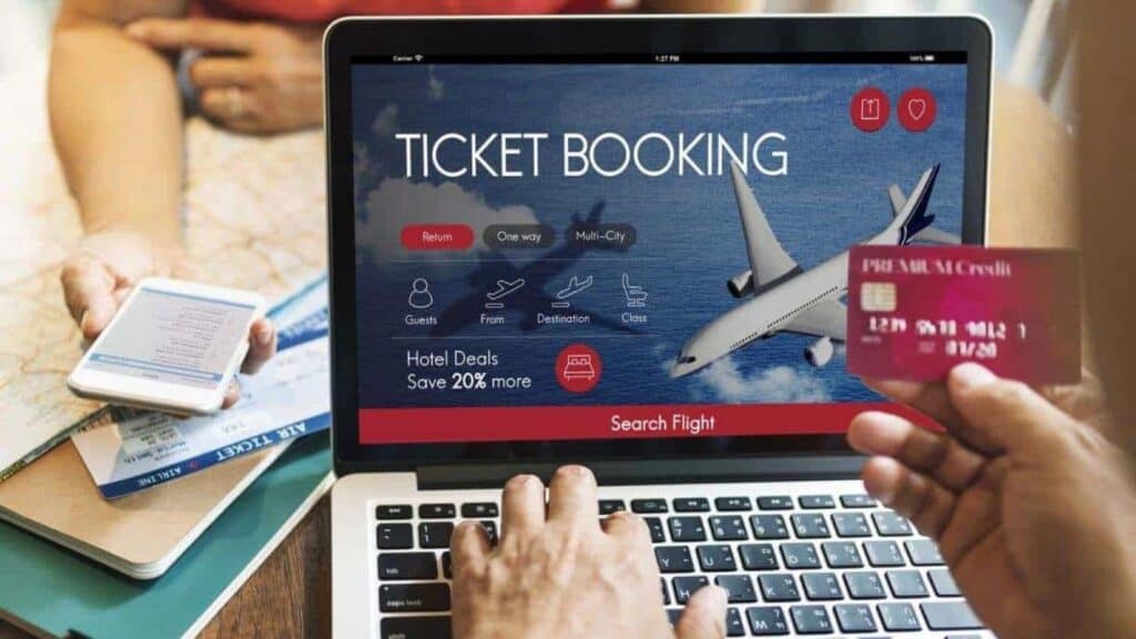 Person booking a flight online using a laptop, with a flight booking website open and airline change and cancellation rules clearly displayed, holding a credit card; another person holds a smartphone and boarding passes.