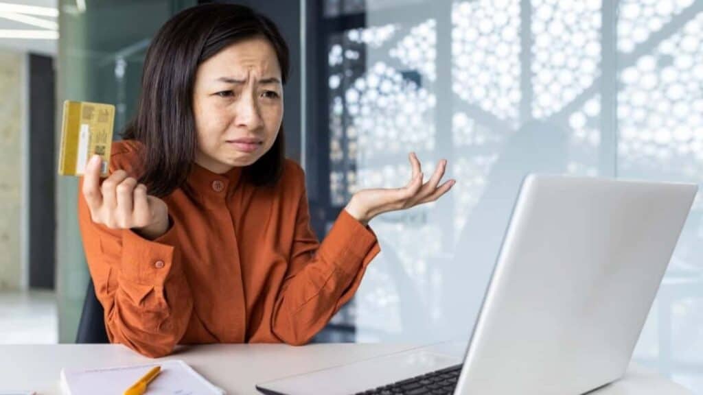 Woman holding a credit card looks confused while sitting at a desk with a laptop, notebook, and pen, possibly trying to understand travel fees before you fly.