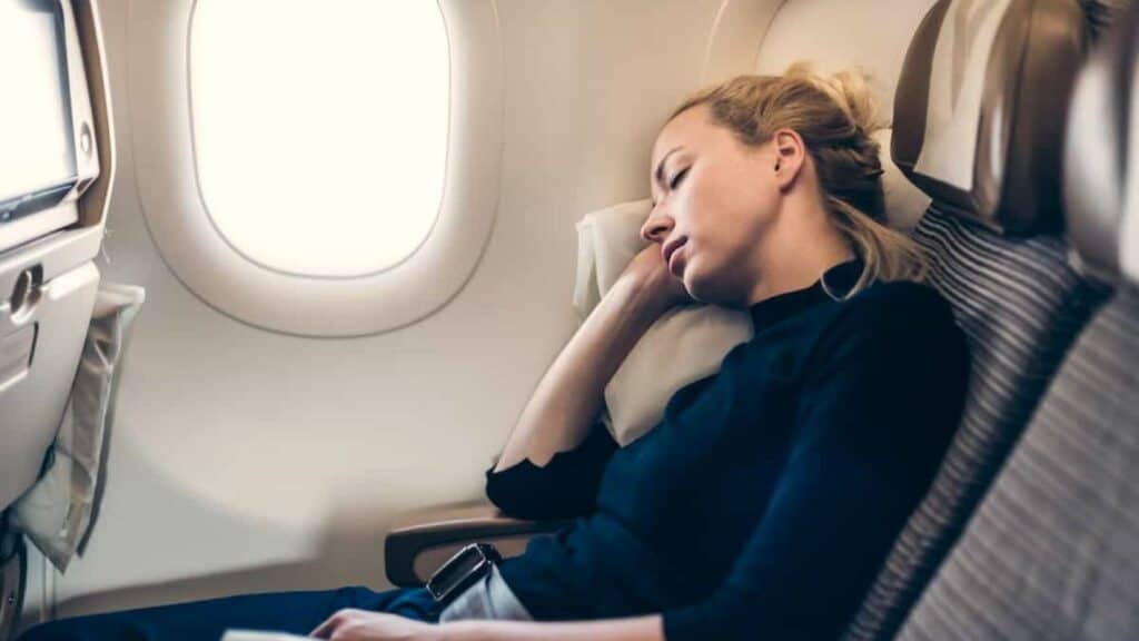 A woman with blonde hair sleeps in her seat on an airplane, leaning on a pillow against the window, mindful of airline basic economy rules that limit her carry-on comforts.