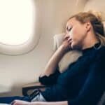 A woman with blonde hair sleeps in her seat on an airplane, leaning on a pillow against the window, mindful of airline basic economy rules that limit her carry-on comforts.