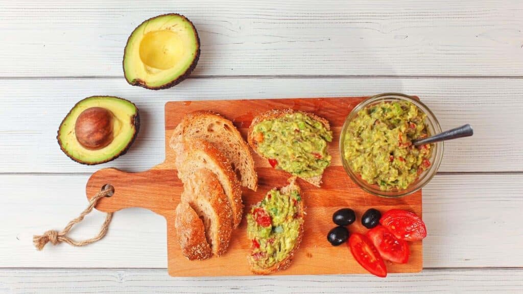 A wooden board with sliced bread, guacamole in a bowl, bread topped with creamy guacamole, black olives, tomato slices, and a halved avocado on a white wooden surface&mdash;perfect for serving as part of your game day dips and spreads lineup.