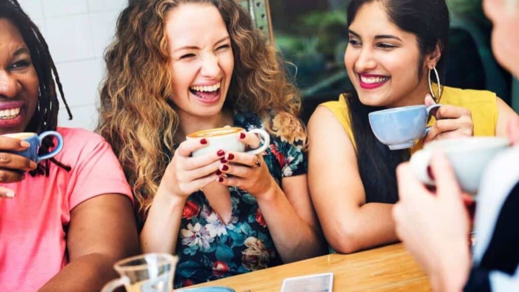 Three women sit at a wooden table, smiling and drinking from mugs, engaged in lively conversation about the states that drink the most coffee. A deck of cards is partially visible on the table.