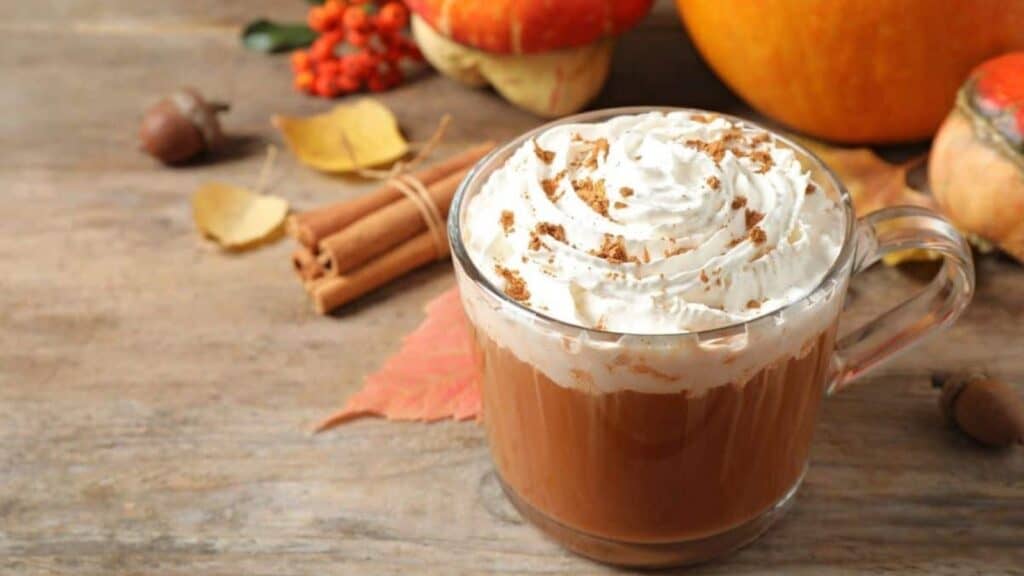 A glass mug of coffee topped with whipped cream and sprinkled cinnamon sits on a wooden table with autumn decorations in the background, capturing the cozy spirit of the Starbucks PSL launch timeline.