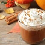 A glass mug of coffee topped with whipped cream and sprinkled cinnamon sits on a wooden table with autumn decorations in the background, capturing the cozy spirit of the Starbucks PSL launch timeline.