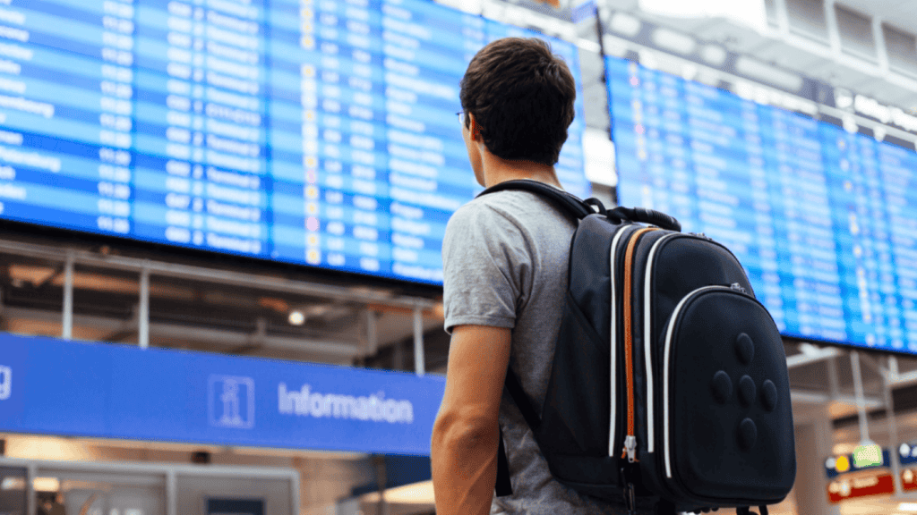 A man with a backpack stands in an airport, looking at a large electronic departures board displaying flight information, carefully reviewing the airline basic economy rules before selecting his flight.