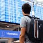 A man with a backpack stands in an airport, looking at a large electronic departures board displaying flight information, carefully reviewing the airline basic economy rules before selecting his flight.