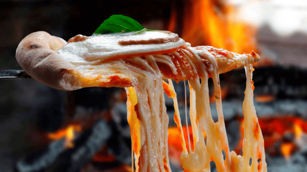 A slice of pizza with melted cheese being lifted from a pizza, with a wood-fired oven and flames visible in the background.