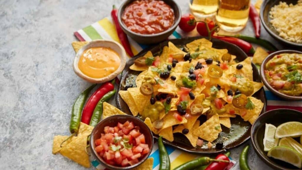 A plate of nachos with cheese, jalape&ntilde;os, and black beans, surrounded by bowls of salsa, guacamole, queso dip, tomatoes, and lime wedges on a striped napkin&mdash;these delicious bites are reminiscent of the standout stadium foods at NFL games.