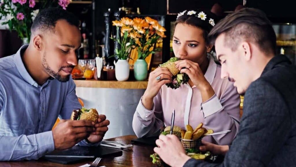 Three people sit at a table in a restaurant, enjoying burgers and fries, with flowers and drinks visible in the background&mdash;a lively scene that reflects the growing trend of US metros dining out spending.