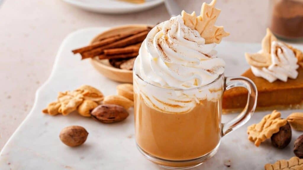 A glass mug of pumpkin spice latte, reminiscent of the cozy excitement around the Starbucks PSL launch timeline, is topped with whipped cream and a leaf-shaped cookie, surrounded by nuts, cookies, and a slice of pie.