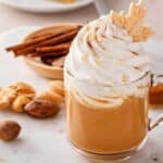 A glass mug of pumpkin spice latte, reminiscent of the cozy excitement around the Starbucks PSL launch timeline, is topped with whipped cream and a leaf-shaped cookie, surrounded by nuts, cookies, and a slice of pie.
