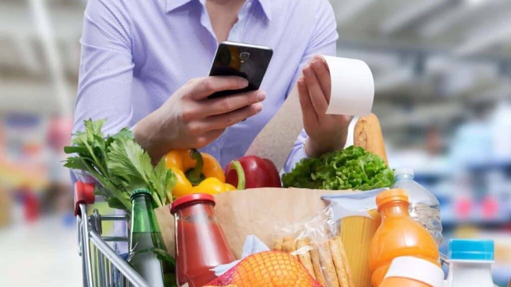Person in a grocery store holding a smartphone and a receipt, with a shopping cart full of various food items—including vegetables, drinks, packaged goods, and plenty of snacks perfect for the Big Game. With Americans expected to eat 1.48 billion wings during the festivities, it’s no wonder this shopper looks ready for game day celebrations.