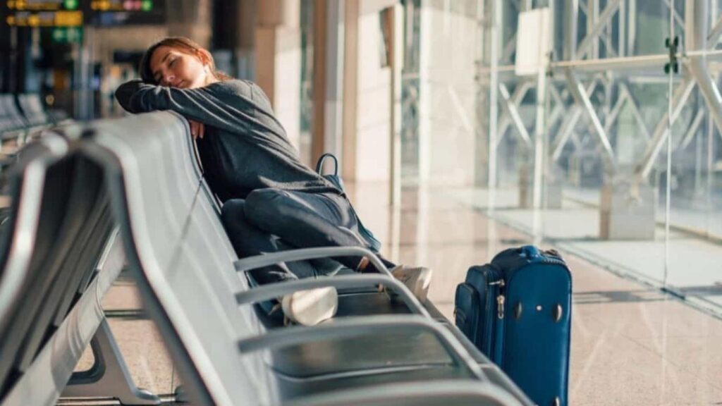 A person is sleeping on airport seats with their arms on the backrest and a blue suitcase placed nearby, likely exhausted from US airports flight delays.