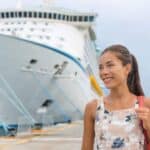 A woman stands on a dock, smiling warmly, with a large cruise ship in the background on a cloudy day—capturing the excitement of cruise ship embarkation and disembarkation as she prepares for her voyage.