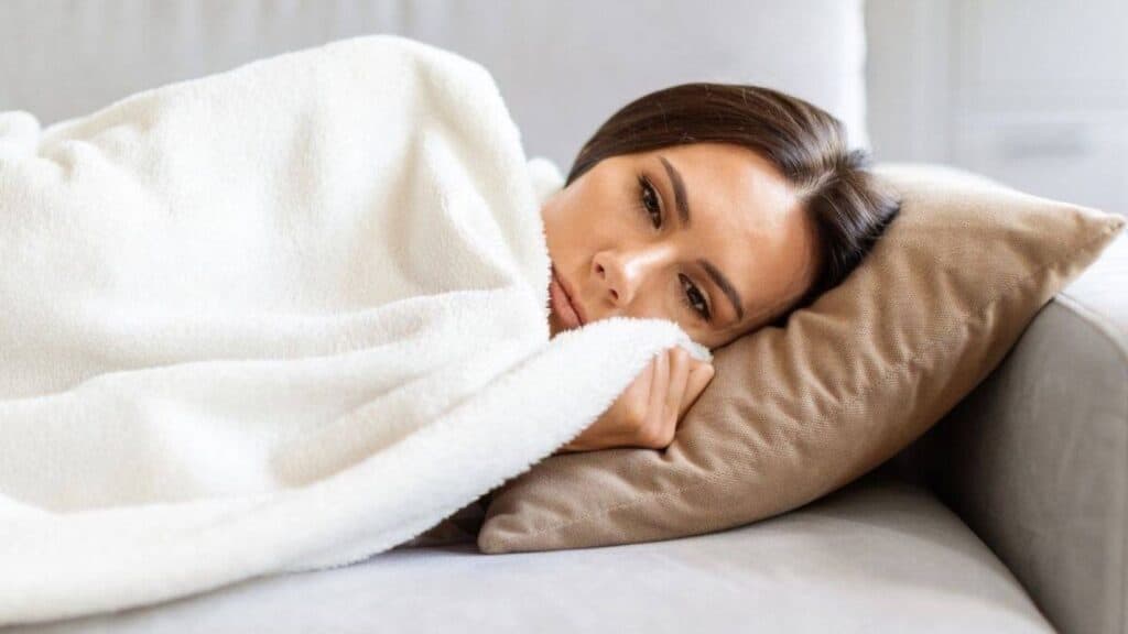 Woman lying on a couch with a beige pillow, covered by a white blanket, looking ahead with a neutral expression—an ideal moment of comfort often found in thoughtful gifts from College Care Package Co.