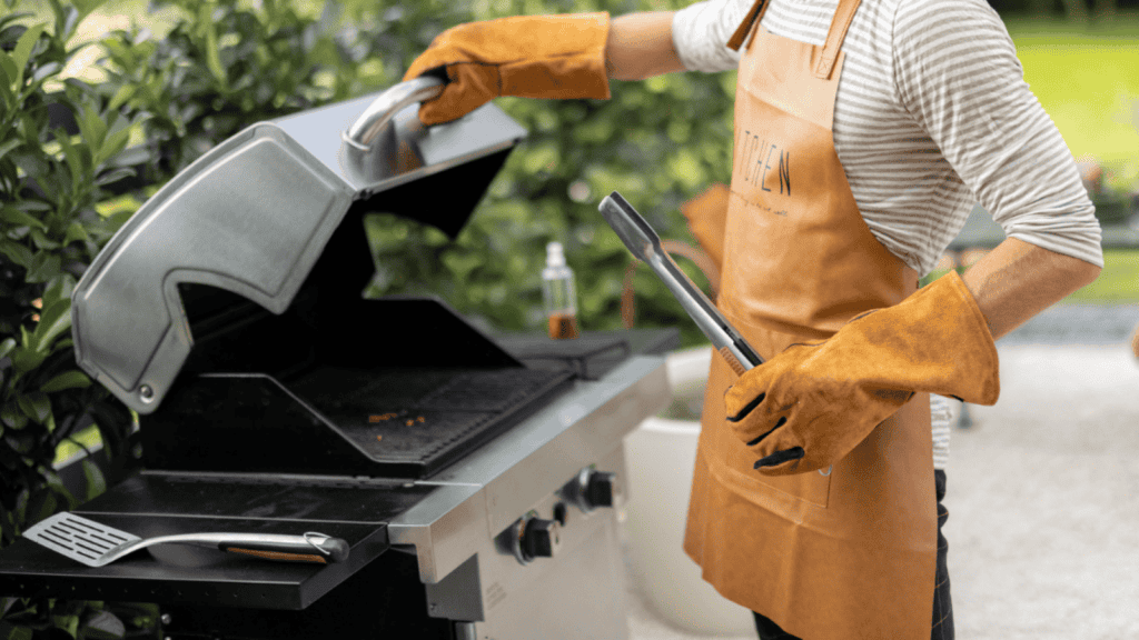 Person wearing gloves and apron opens a grill with one hand and holds tongs in the other. A spatula is on the grill and plants are visible in the background, highlighting how premium tailgating gear elevates even simple outdoor moments into a luxury category.