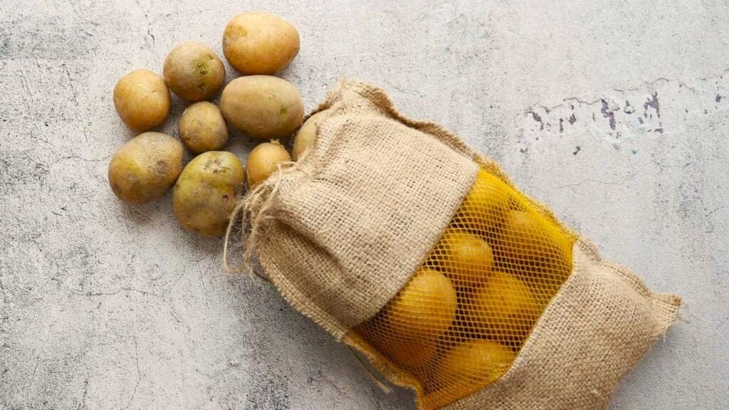 Several potatoes, classic fall vegetables coming into season, spill out of a burlap sack with a yellow mesh window, resting on a light gray textured surface.