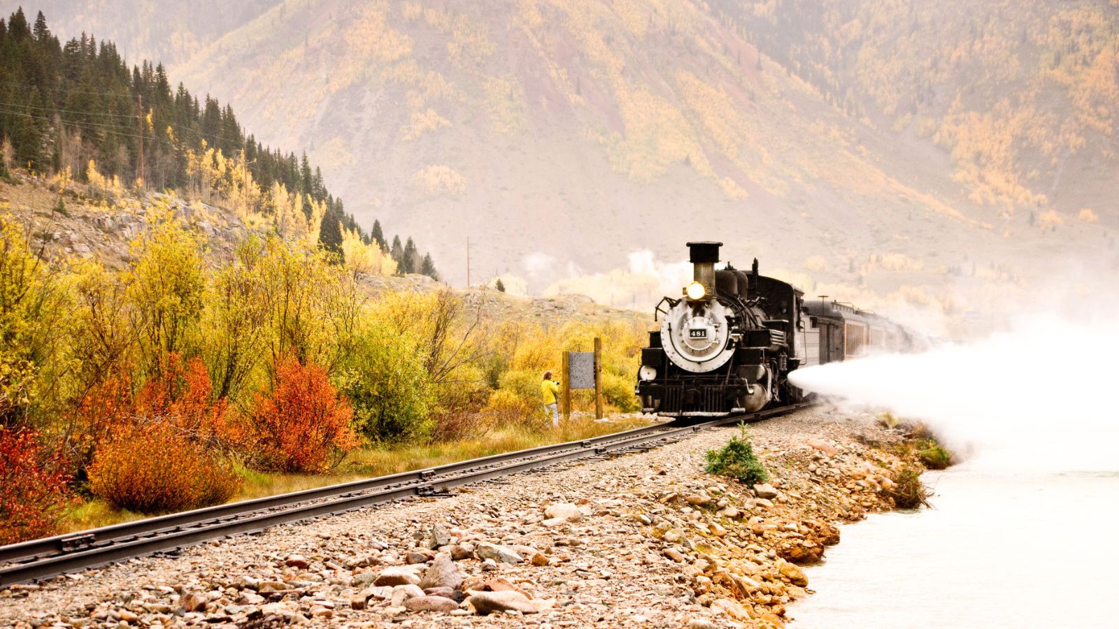 A steam locomotive travels along a railway track beside a river, with autumn trees and mountains in the background. Steam billows from the train.