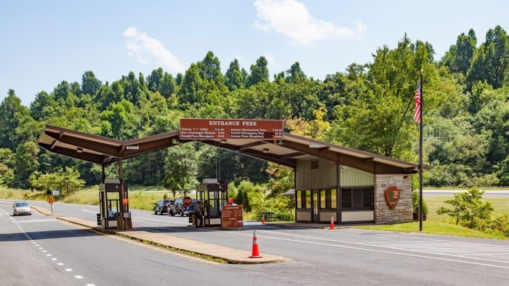 A park entrance station with two covered lanes, fee signs noting upcoming national park fee free days, and a guard booth. Vehicles wait in line under clear skies, surrounded by trees and greenery.