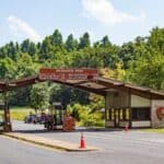 A park entrance station with two covered lanes, fee signs noting upcoming national park fee free days, and a guard booth. Vehicles wait in line under clear skies, surrounded by trees and greenery.