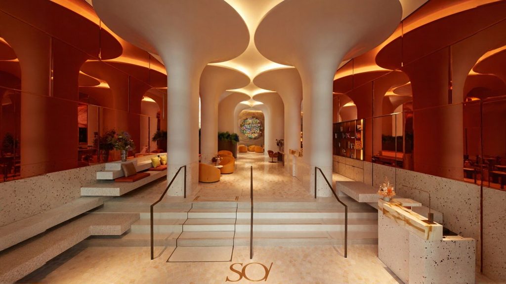 Modern interior with sculptural white columns reminiscent of the Eiffel Tower, warm lighting, terrazzo stairs, and a seating area with orange accents. The floor features the letters “SOY” near the entrance.