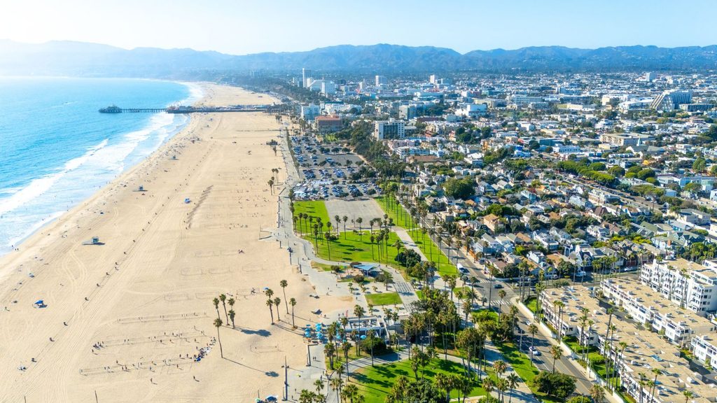 Aerial view of a wide sandy beach, adjacent park with palm trees, parking lots, and a coastal city with mountains in the background under a clear sky.