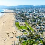 Aerial view of a wide sandy beach, adjacent park with palm trees, parking lots, and a coastal city with mountains in the background under a clear sky.