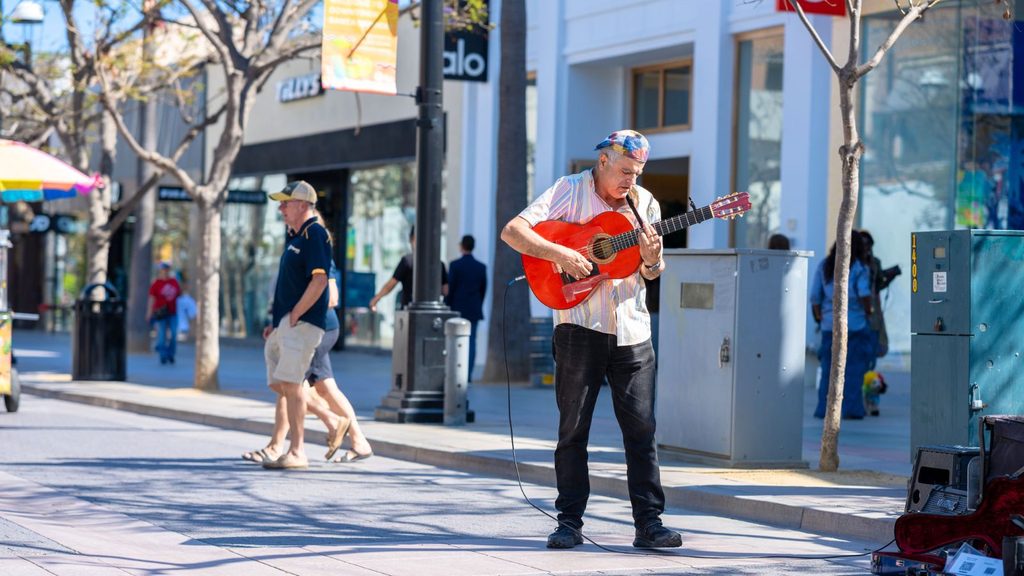 A man plays a red acoustic guitar on a city sidewalk while pedestrians walk by. Shops and trees line the street in the background.