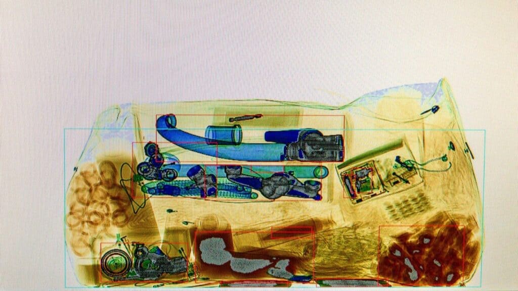 X-ray image of a bag showing various objects, including mechanical parts, wires, circular items, and rectangular components arranged within the bag&mdash;a typical view during TSA liquid screening at US airports.
