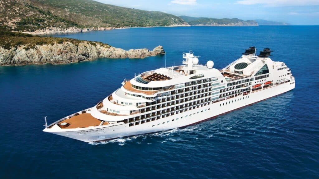 A large white cruise ship sails near a rocky coastline with green hills under a clear blue sky.