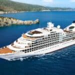 A large white cruise ship sails near a rocky coastline with green hills under a clear blue sky.