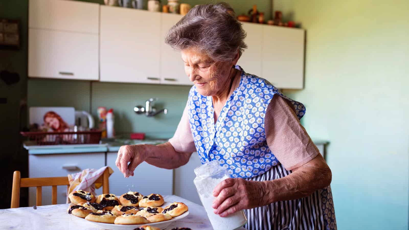 An elderly woman in an apron sprinkles sugar from a jar onto pastries in a kitchen with white cabinets and a green wall.