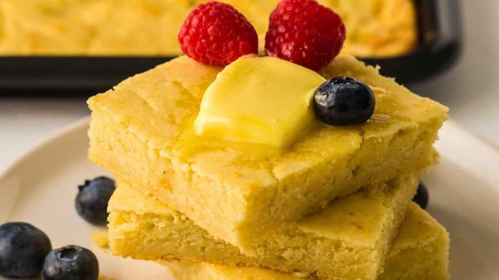 Three pieces of pancakes on a plate, topped with a pat of butter, two raspberries, and a blueberry, with extra blueberries nearby.