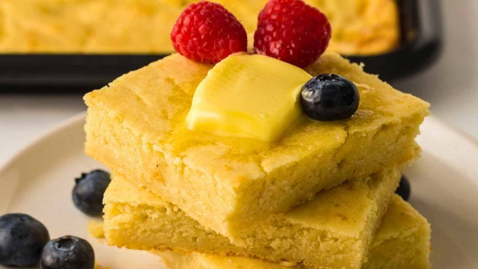 Three pieces of pancakes on a plate, topped with a pat of butter, two raspberries, and a blueberry, with extra blueberries nearby.