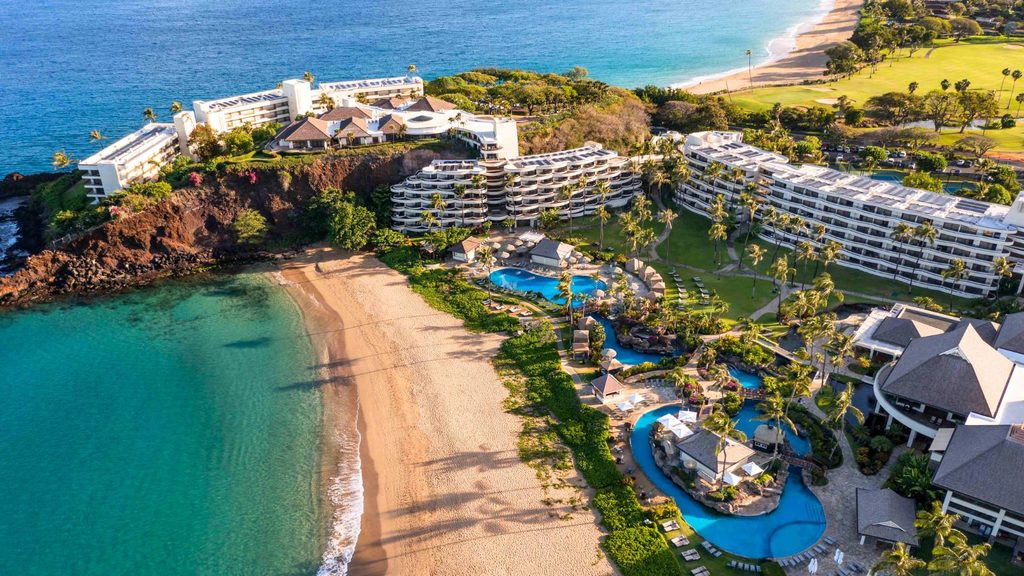 Aerial view of a beachfront resort with multiple buildings, swimming pools, palm trees, and direct access to a sandy beach by clear blue water.