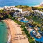 Aerial view of a beachfront resort with multiple buildings, swimming pools, palm trees, and direct access to a sandy beach by clear blue water.