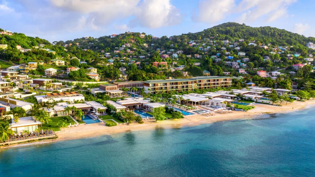 Aerial view of a beachfront resort with pools, buildings, and palm trees, bordered by hills with houses and lush greenery.