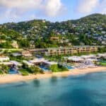 Aerial view of a beachfront resort with pools, buildings, and palm trees, bordered by hills with houses and lush greenery.