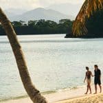 Two people walk along a sandy beach near calm water, with palm trees in the foreground and mountains in the background.