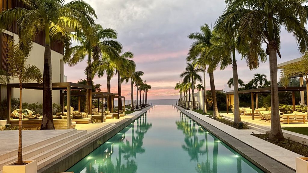 Infinity pool lined with palm trees and lounge seating, leading toward the ocean at sunset under a partly cloudy sky.