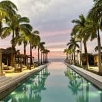 Infinity pool lined with palm trees and lounge seating, leading toward the ocean at sunset under a partly cloudy sky.